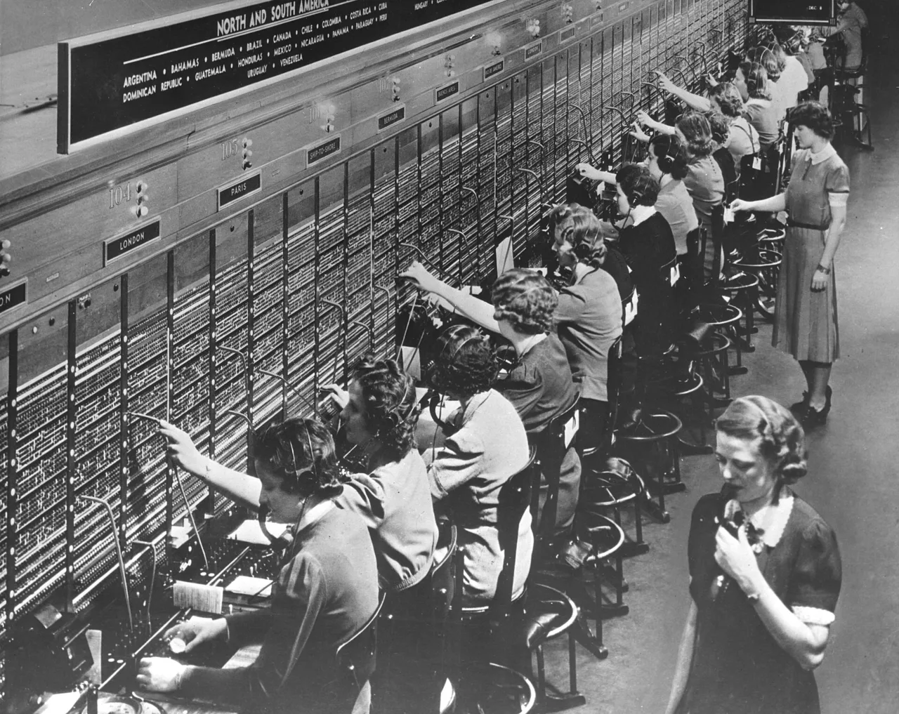 Архивный снимок телефонной станции Bell System: ряд операторов в наушниках соединяет линии на коммутаторе с табличками городов